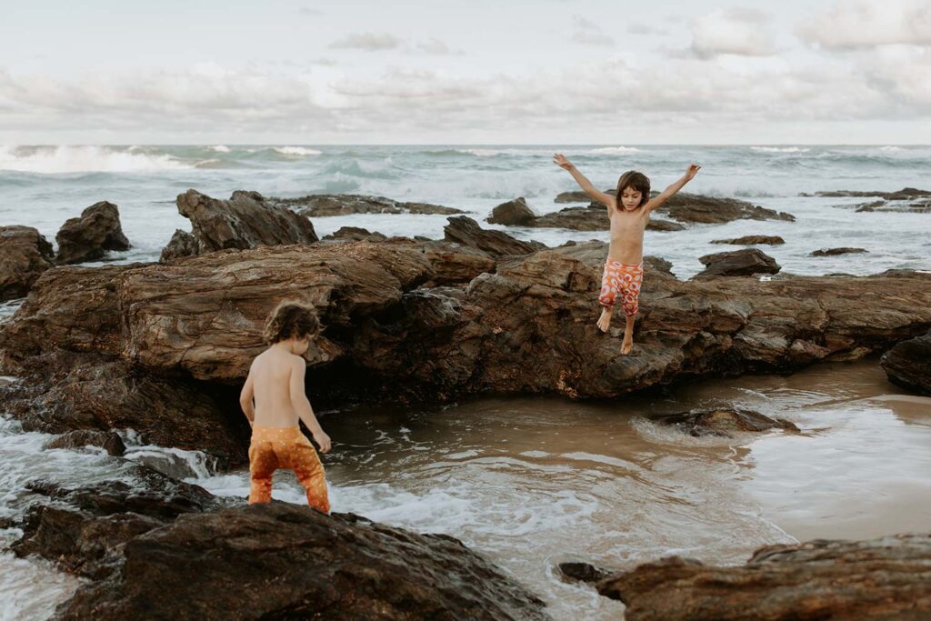 two young brothers are playing on the rocks at the beach and one is about to jump in the water at family photo shoot