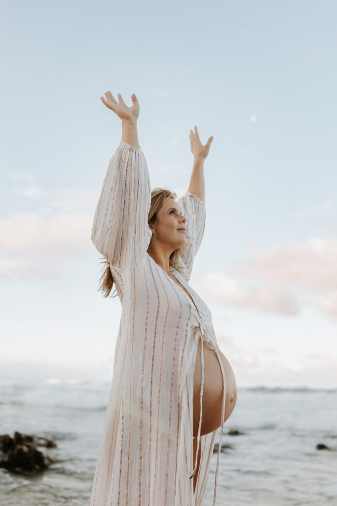 pregnant woman at the beach she has her arms in the air smiles and her belly is showing