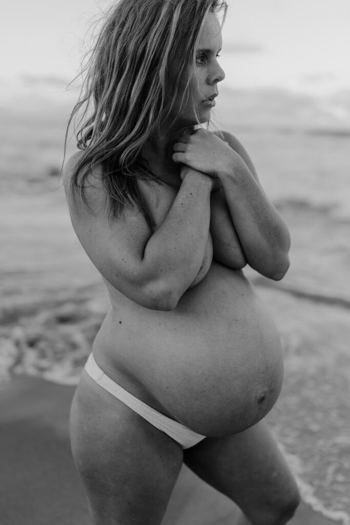 black and white image of a pregnant woman she is only wearing her underwear and covering her breasts with her arms at the maternity photoshoot