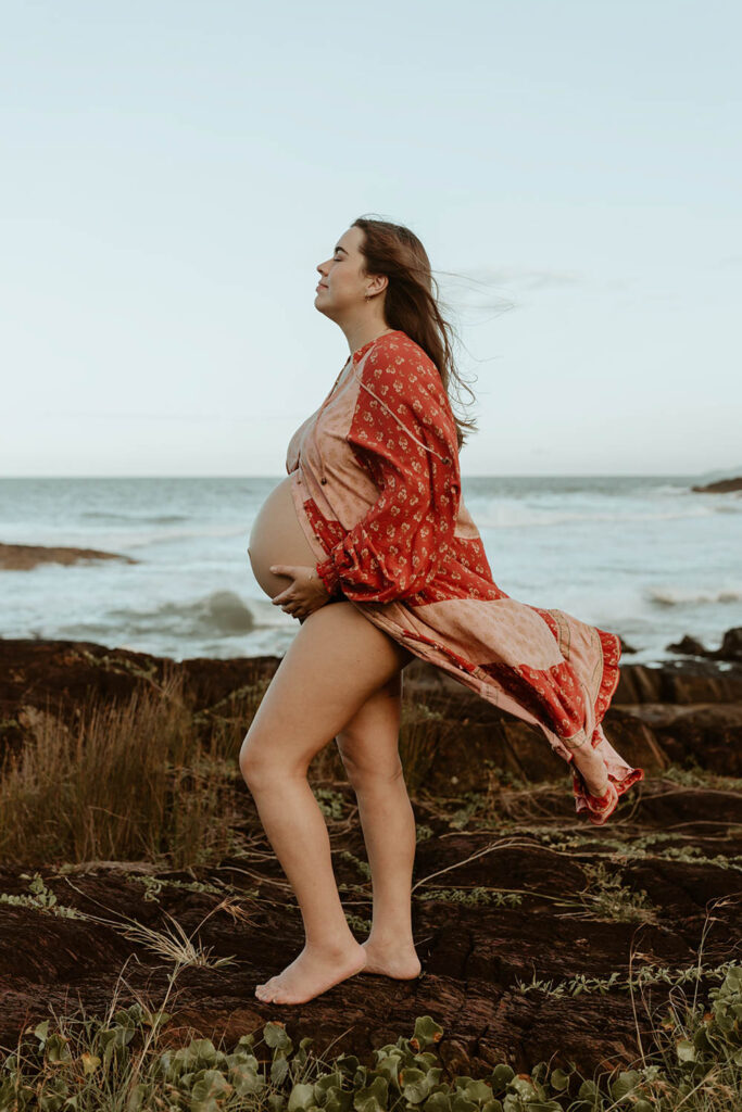 pregnant woman at the beach standing on a rock she holds her belly and has a eyes closed