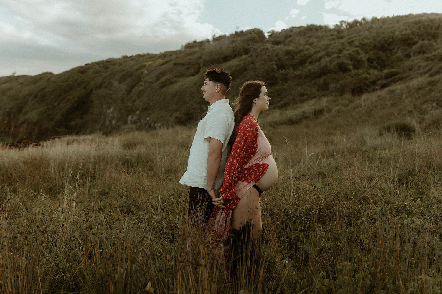 pregnant couple standing back to back in a field with long grass