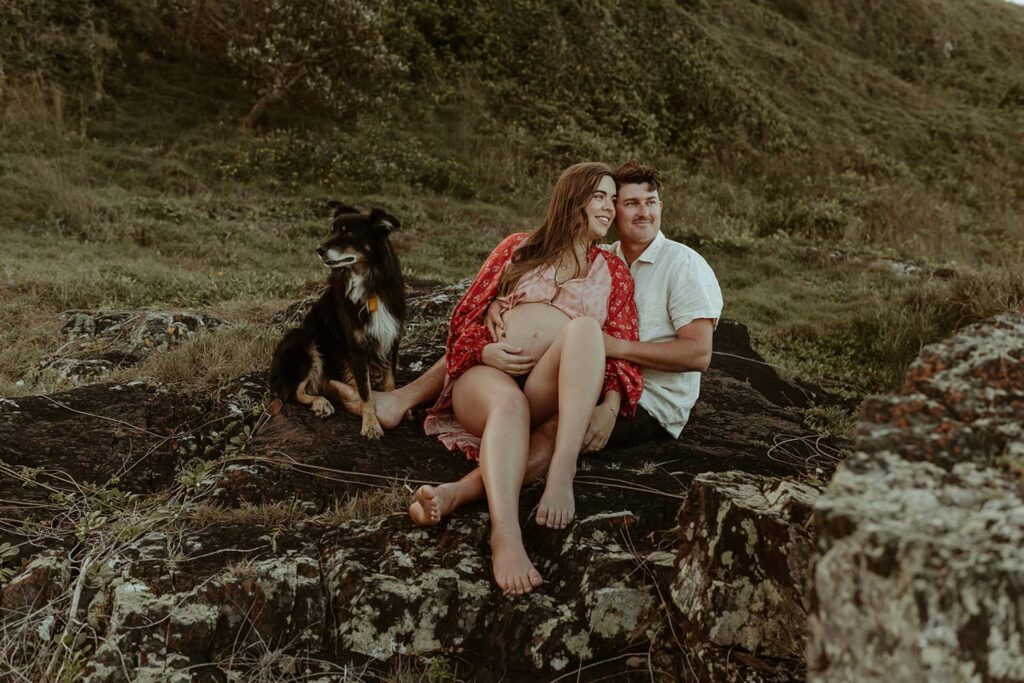 pregnant couple seated on a rock with their dog near them