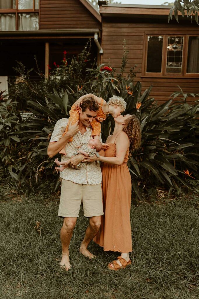 A family is standing outside in front of their house  the dad is holding their newborn baby and also carry  their toddler on his shoulder the toddler is kissing his mum