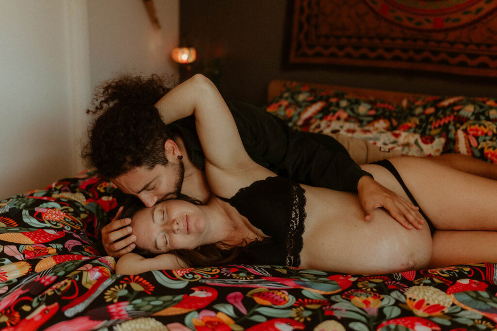 A pregnant couple lying on their bed snuggling, the man is holding the pregnant belly while kissing his wife on the head